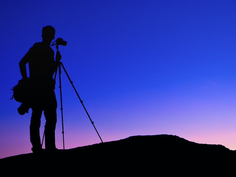 Silhouette of a photographer and tripod standing on top of a hill. It is sunset or sunrise and the sky is clear with a bold colour gradient from blue to violet to yellow.